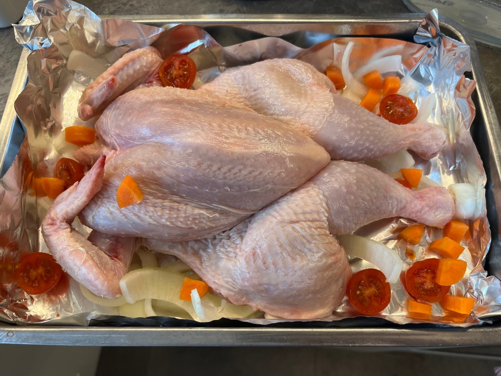 Raw spatchcocked chicken placed in a roasting dish with sliced onions, cherry tomatoes, and diced carrots on a bed of foil.