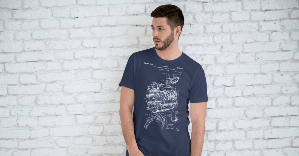 White, male model wearing a blue T shirt with Frank Whittle's jet engine design. He is standing in front of a white painted brick wall.