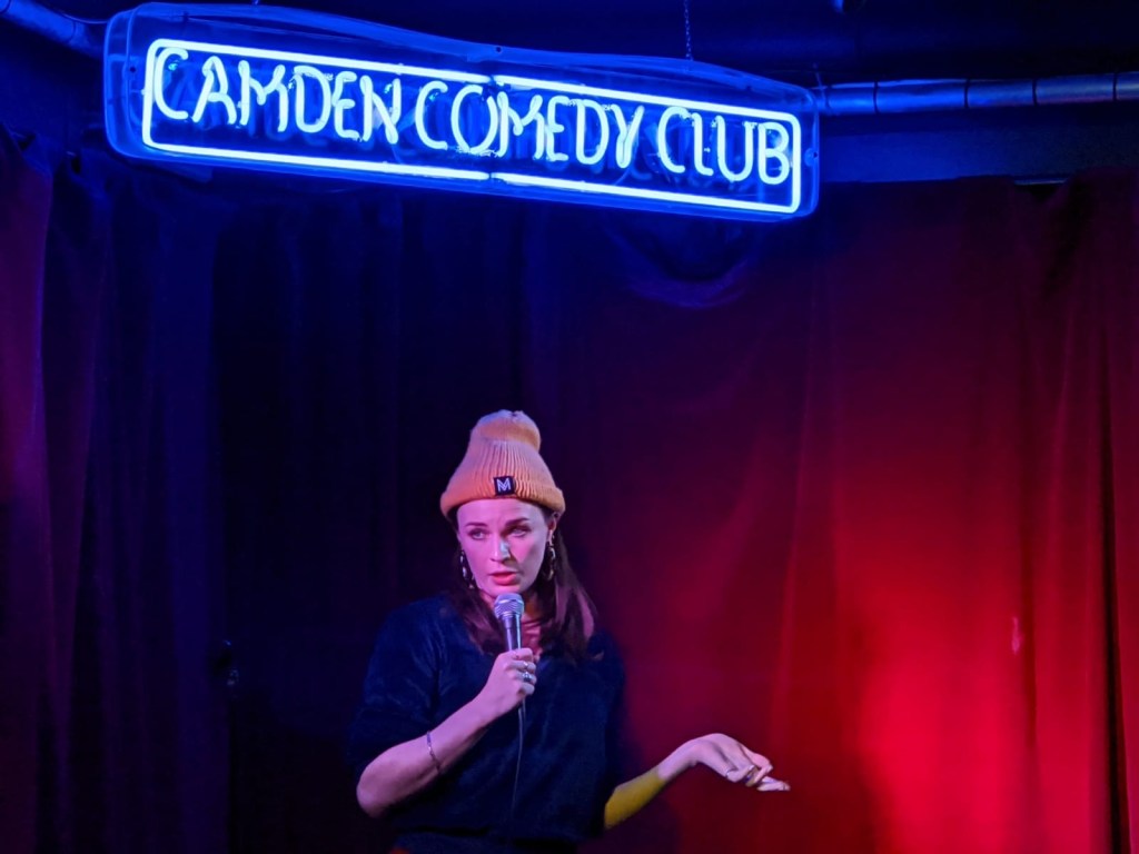 Aisling Bea wearing an orange hat under a neon sign reading 'Camden Comedy Club'. 